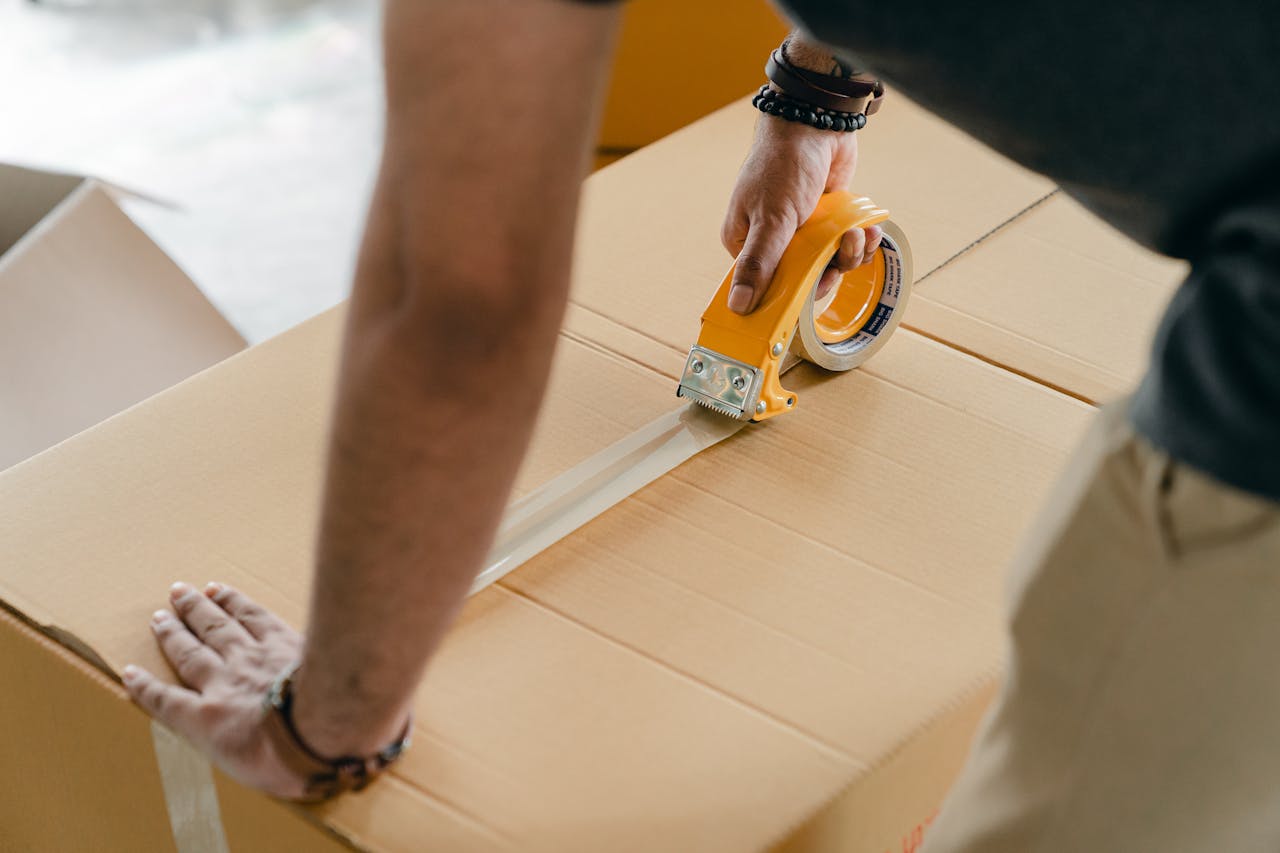 about-us-01 Man using packing tape to seal a cardboard box indoors. Ideal for moving or shipping concepts.