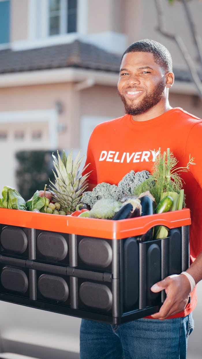 services-02 A deliveryman in a red shirt delivering a crate of fresh vegetables outdoors.