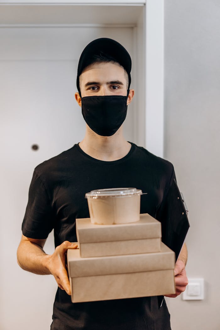 Mastering the First Impression: Your intriguing post title goes here A young delivery man in a black mask holds food boxes indoors, ready for delivery.